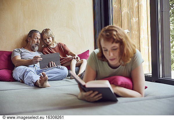Father and daughter using digital tablet while woman reading book on bed at home