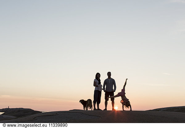 Father and mother looking at girl performing cartwheel on rock formation against sky during sunset