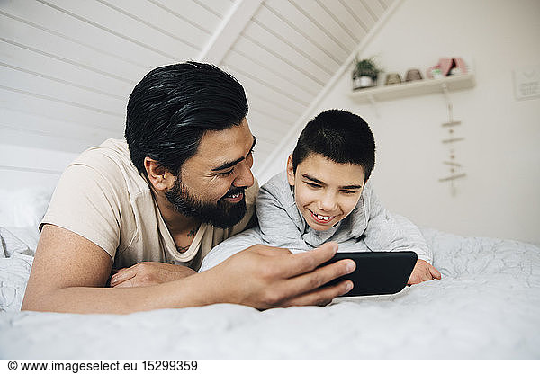 Father and smiling disabled son watching movie over mobile phone while lying on bed at home