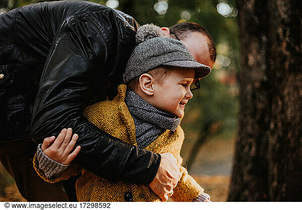 Father and son having hug in autumn city park. Happy kid walking among fallen leaves. Kids fashion. Boy wearing trendy yellow coat  cap and scarf. Smiling young boy outdoors. Kid jumping and