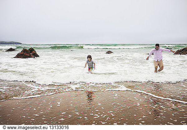 Father and son playing on waves at shore against cloudy sky