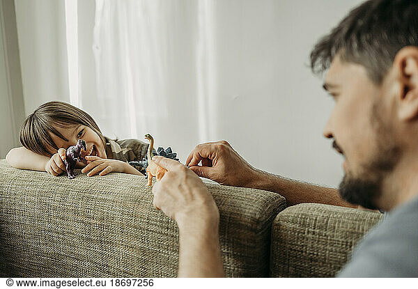 Father and son playing with toy dinosaurs on sofa at home