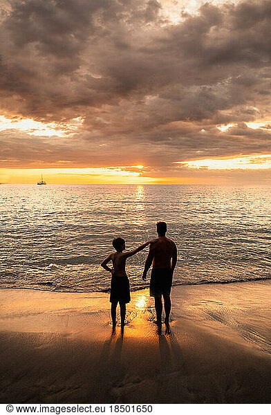 Father and son standing on a sandy beach watching the sunset together.