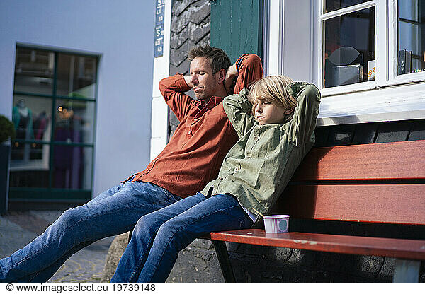 Father and son with hands behind head sitting on bench