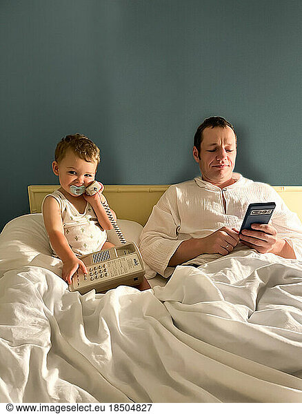 Father and son with pacifier hold telephone on bed