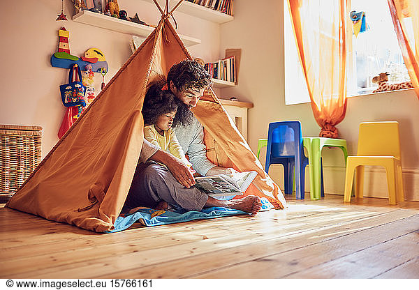 Father and toddler daughter reading book in teepee