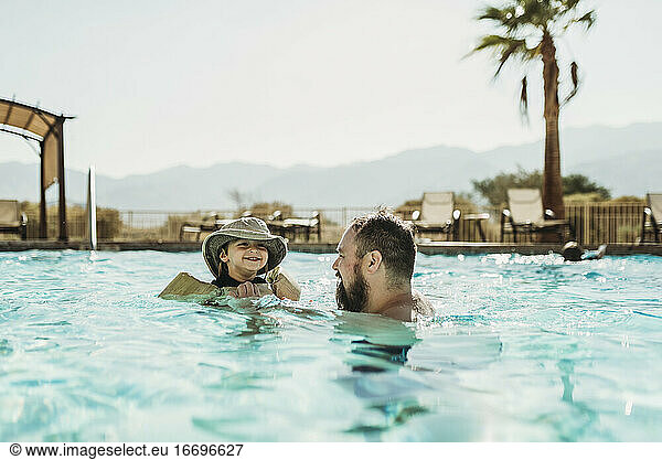 Father and toddler son swimming in pool with mountains and palm trees