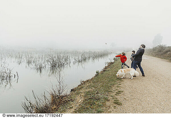 Father and two sons with dog throwing stones into lake on foggy day
