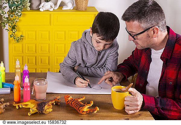 Father assisting his son in drawing while having a coffee.