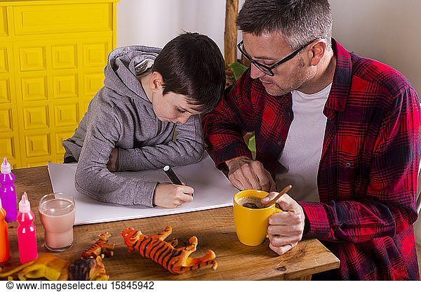 Father assisting his son in drawing while having a coffee.