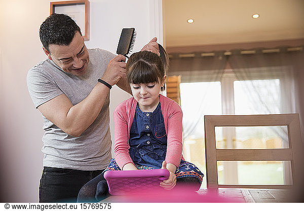 Father brushing hair of daughter using digital tablet