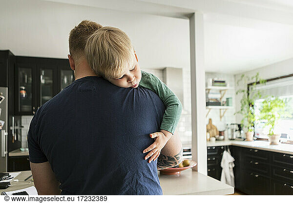 Father carrying sleeping son in kitchen