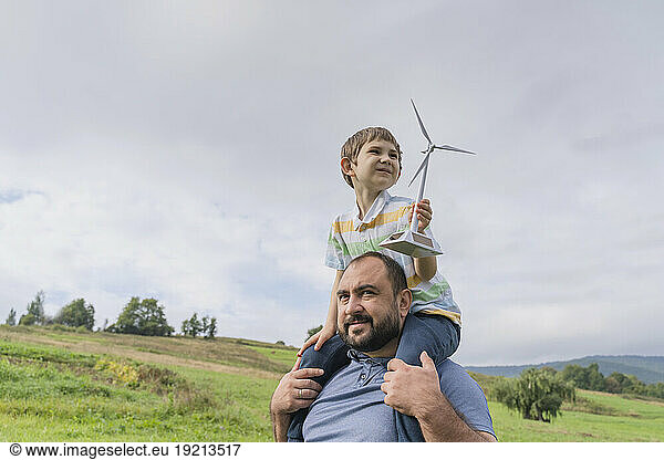Father carrying son on shoulders with wind turbine model in front of sky