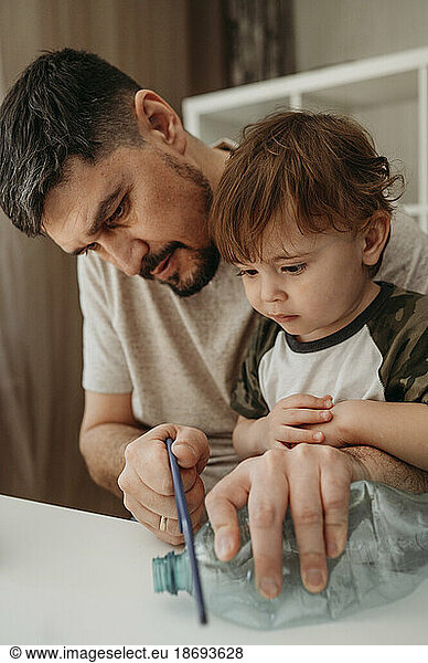 Father cutting plastic bottle with hand saw by son at home