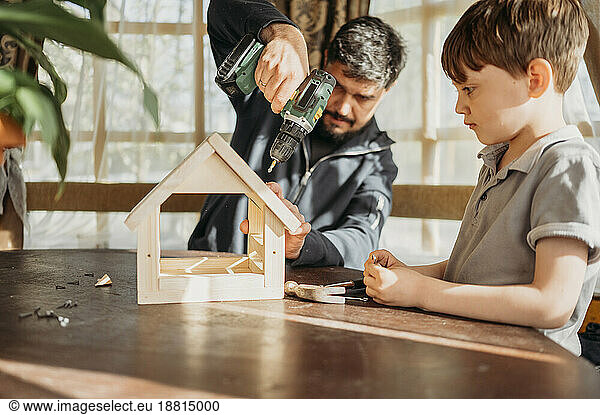 Father drilling birdhouse on table with son at home