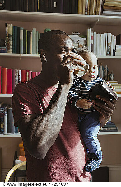 Father drinking coffee while carrying male toddler at home