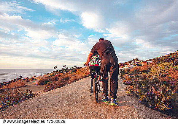 Father helping son ride his bike on a coastal trail.