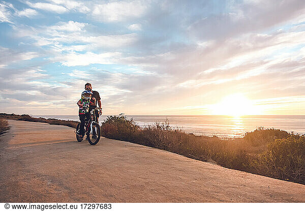 Father helping son ride his bike on a coastal trail at sunset.