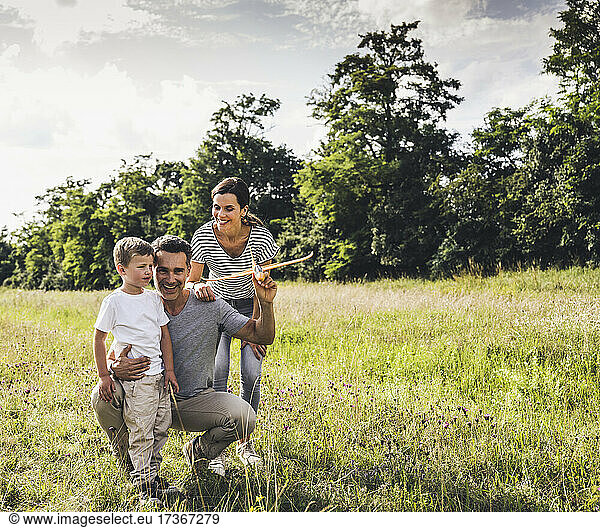 Father holding airplane toy while crouching by son and woman on grass