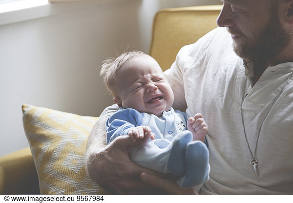 Father holding and looking at crying baby  sitting on sofa