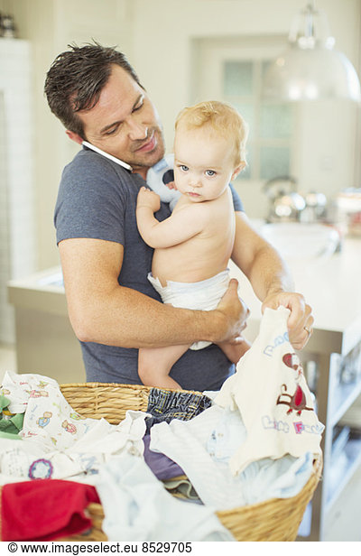Father holding baby and folding laundry while talking on cell phone