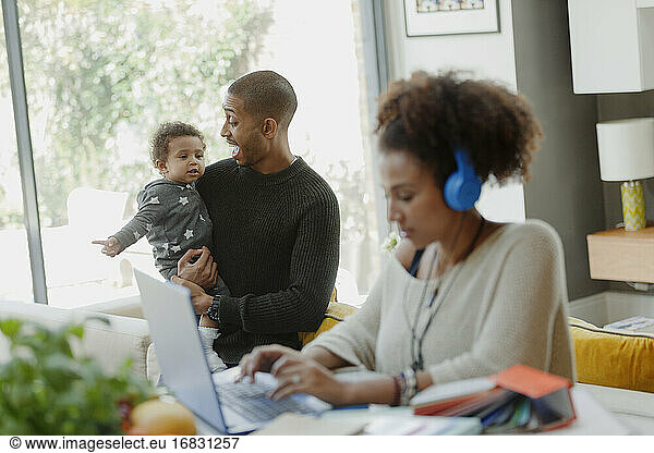 Father holding baby daughter behind working mother at laptop
