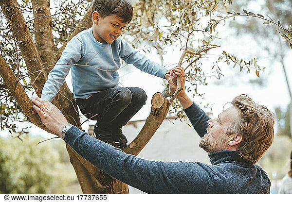 Father holding hands of son crouching on tree