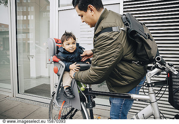 Father holding son sitting in safety seat of bicycle on sidewalk in city