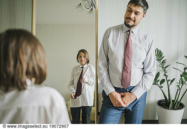 Father looking at son trying necktie in front of mirror at home