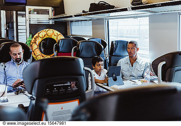 Father looking at son using digital tablet and businessman working at desk in train