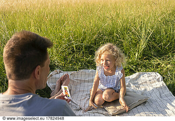 Father playing card with cheerful daughter sitting on picnic blanket