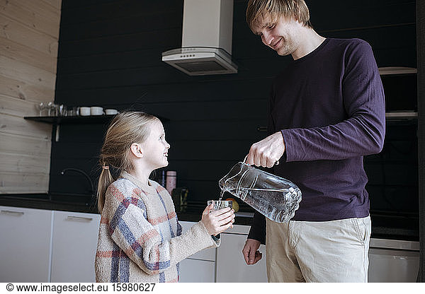 Father pouring water into glass for his smiling daughter in the kitchen
