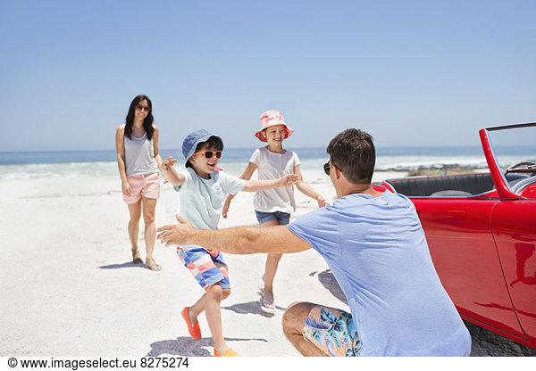 Father reaching to hug children on beach next to convertible