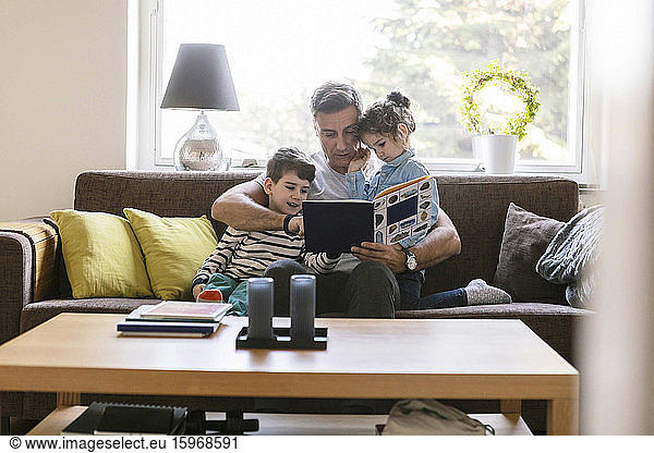 Father reading book to son and daughter while sitting on sofa at home