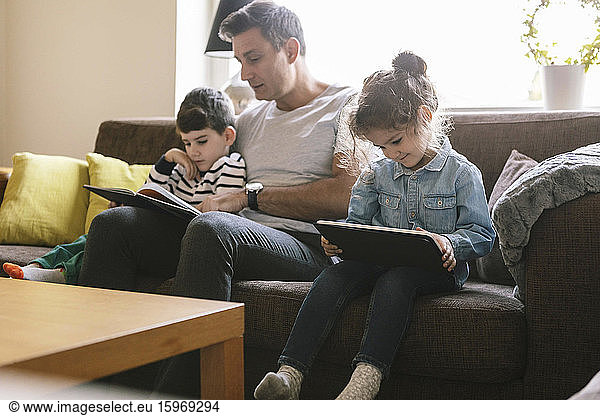 Father reading book to son while daughter using digital tablet on sofa at home