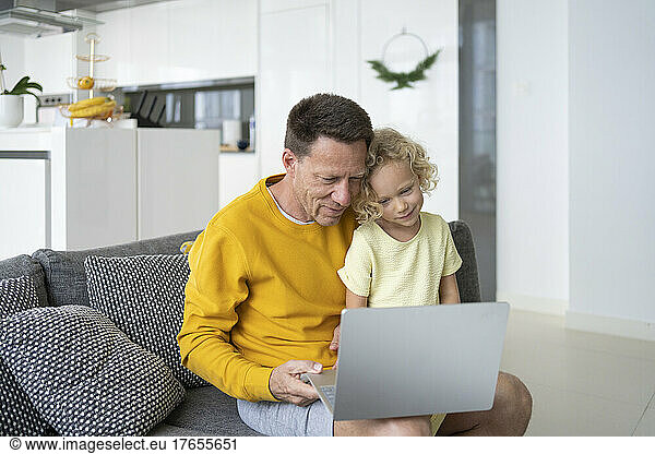 Father sharing laptop with daughter sitting on sofa at home