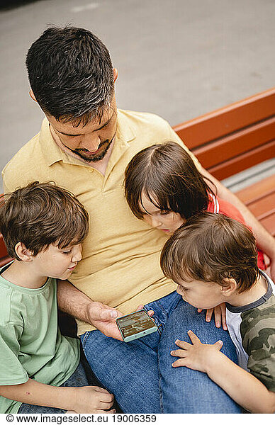 Father sharing smart phone with sons sitting on bench at park