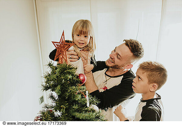 Father showing Christmas tree decoration while carrying daughter by son against wall at home