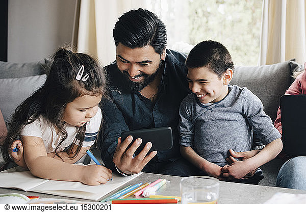 Father showing mobile phone to children while woman using laptop on sofa in living room