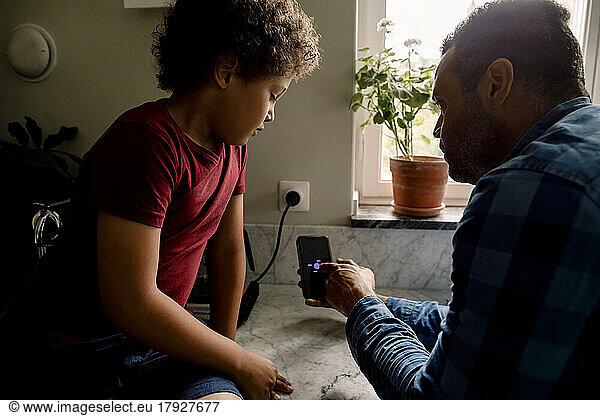 Father showing smart phone screen to son sitting on kitchen counter at home