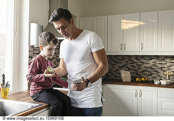 Father showing smart phone to son with food while standing by kitchen counter