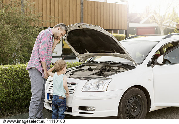 Father talking on phone and looking at car engine while standing by boy on street