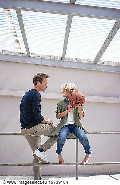 Father talking to son holding basketball and sitting on railing