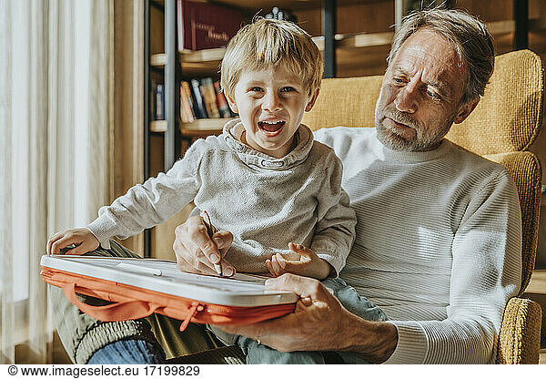 Father teaching drawing to cheerful son while sitting on chair