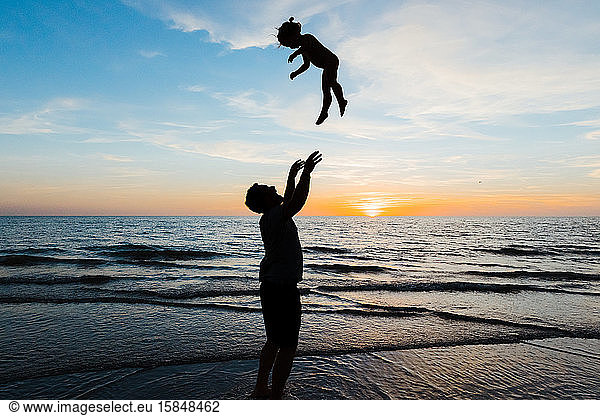 father throws his daughter into the air at sunset on beach vacation