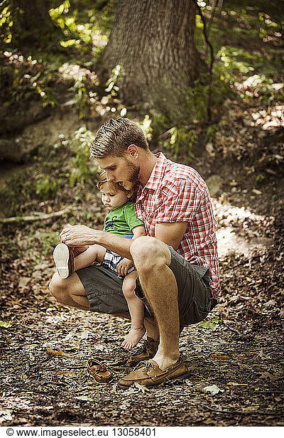 Father tying son's shoelace while crouching on field in forest