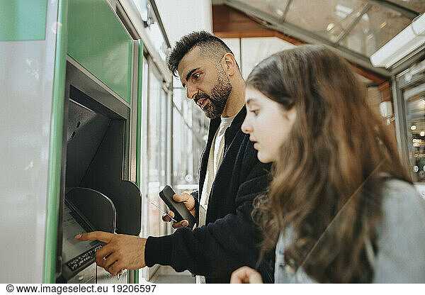 Father using ATM machine while standing with daughter at railroad station
