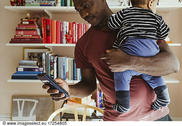 Father using smart phone while carrying baby boy at home