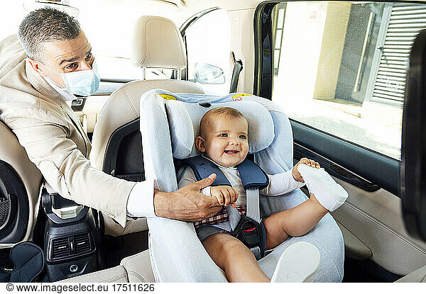 Father wearing protective mask baby boy sitting in child's seat in a car