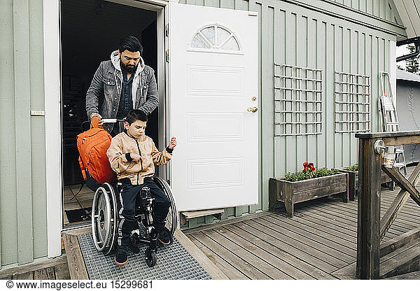 Father with backpack pushing autistic son sitting on wheelchair at doorway